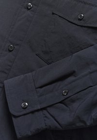 Camisa de manga larga azul oscura con puños abotonados, un bolsillo frontal con tapa abotonada y detalles de costura visibles.