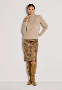 Frau in beigem Rippstrickpullover, gemustertem knielangem Rock und hellbraunen Wildlederschuhen bis zum Knie, die vor einer schlichten hellen Wand steht.