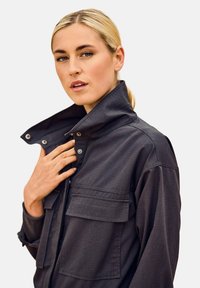 Donkergrijze jas met een hoge kraag, twee voorkruiszakken en drukknoopdetails, met een subtiel textuurpatroon over de gehele jas.