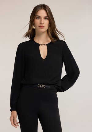 Mujer con cabello castaño lleva una blusa negra de manga larga con escote en forma de ojo de cerradura y detalle de cadena dorada, combinada con pantalones negros y cinturón a juego.