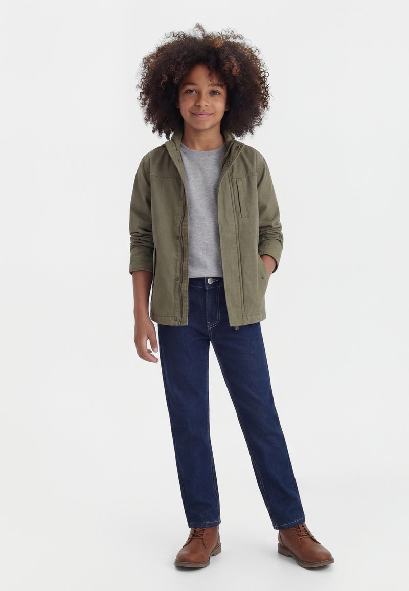 Barn med krøllet hår iført en olivengrøn jakke, grå skjorte, mørkeblå jeans og brune støvler, står mod en hvid baggrund.