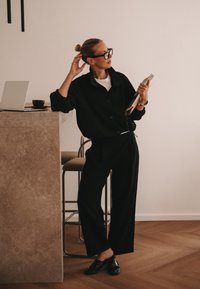 Woman in black outfit and glasses leans on counter holding a folded magazine, with laptop and coffee cup on the counter behind her.