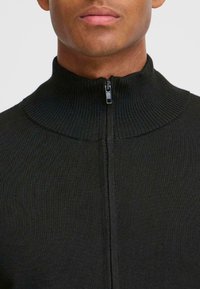 Närbild av en persons nacke och haka iklädd en mörkstickad hög krage med dragkedja. Neutral bakgrund framhäver plagget.