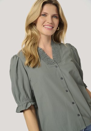 Blouse vert clair avec des manches courtes bouffantes, présentant un col smocké et de petits boutons à l'avant. Tissu doux avec une coupe décontractée.