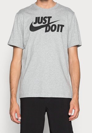 Mężczyzna w szarej koszulce z czarnym napisem "JUST DO IT" i logo Nike, w czarnych spodniach, stojący na jednolitym tle.