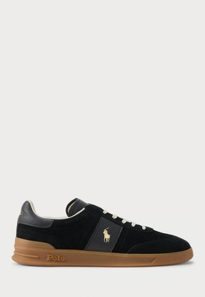 Sneaker in suede nero con dettagli in pelle, lacci bianchi e suola in gomma naturale. Presenta un logo dorato di lato e branding sul tallone.