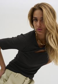 Zwart geribbeld topje met korte mouwen, voorzien van een uitsnijding bij de halslijn, gecombineerd met een gouden ketting en lichtbeige broek.