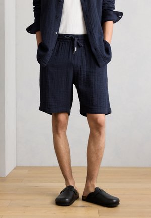 Mężczyzna w granatowych teksturowanych szortach z troczkami, pasującej otwartej koszuli, białym topie i czarnych wsuwanych chodakach stoi na drewnianej podłodze.