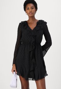 Vestido negro con lunares, mangas largas y transparentes, cuello y dobladillo con volantes, cintura ajustada, textura detallada, combinado con un bolso de color púrpura claro.