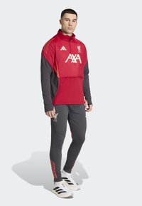 Red tracksuit top with grey sleeves, featuring a high collar and Liverpool FC logo. Paired with grey pants and black and white footwear.