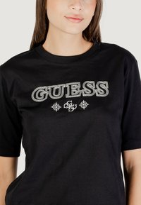 T-shirt in cotone nero con un grande logo ricamato bianco "GUESS" e simboli decorativi sottostanti. Maniche corte e scollo rotondo.