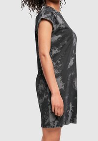 Femme aux cheveux bouclés portant une robe courte à manches courtes tie-dye noire et grise, vue de profil sur un fond clair uni.