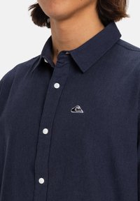 Camisa de botones azul marino en tejido texturizado, que presenta un cuello puntiagudo y un pequeño logo bordado en blanco en el pecho.