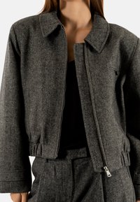 Grijze visgraatjacket met een kraag hals, elastische taille en ritssluiting. Stof lijkt gestructureerd met een textuurdesign.