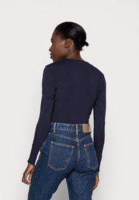 Marineblå langærmet top parret med højtaljede blå jeans, der har gul syning og en læder bagpatch. Glat stofstruktur synlig.