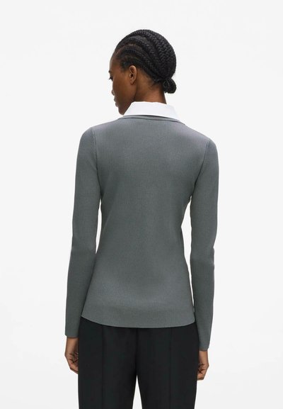 Femme aux cheveux tressés portant un pull gris à manches longues en côte et un pantalon noir, vue de dos sur un fond blanc uni.
