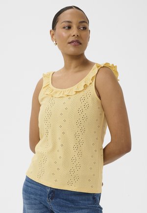 Kvinde iført gul ærmeløs bluse med hulmønster og flæsekant ved halsen samt blå jeans, kigger til siden mod en ensfarvet baggrund.