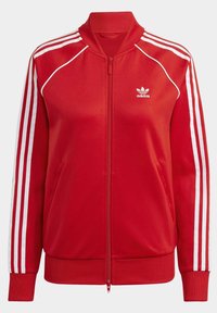 Červená bunda na zip vyrobená z hladké látky, s bílými pruhy podél rukávů a logem Adidas na levé straně hrudi.