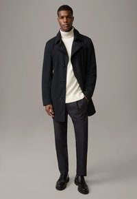 Zwarte overjas met knoopsluiting, gedragen over een witte coltrui en donkere broek. Soepele textuur met strakke, modern lijnen.