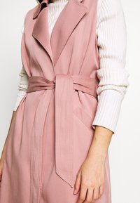 Růžová vesta s páskem a na zakázku šitým designem, vyrobená z lehké látky. Má klopový límec a boční kapsy. Viditelný strukturovaný žebrovaný svetr.