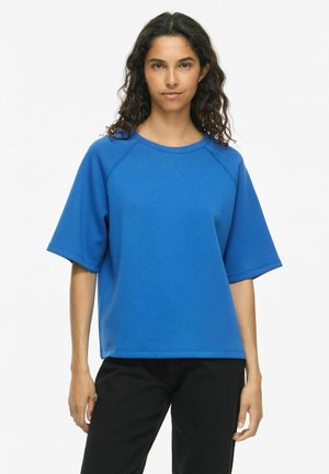 Donna con capelli ricci scuri che indossa una maglia blu a maniche corte larga e pantaloni neri su uno sfondo bianco semplice.