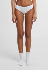 Baltas bikini apakšbikses ar gludu apdari, zemu jostasvietu un minimālistisku dizainu, kopā ar baltām volāna raksta potīšu zeķēm uz kailām kājām.