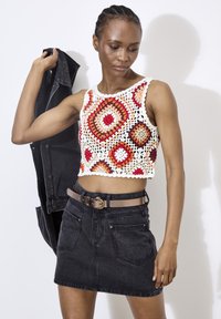 Haut au crochet avec des motifs hexagonaux multicolores en rouge, orange et beige ; associé à une jupe en denim noire et une ceinture. Blouson noir drapé sur l'épaule.