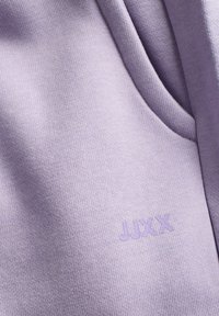 Lilla stof med en glat tekstur, der fremviser et syet logo "JUXX" i samme farve. Detaljer af stofvævningen er synlige.