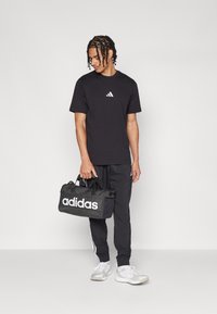 Fekete pamut póló kis fehér Adidas logóval, fekete tréningnadrággal, fehér oldalsávokkal és fehér sportcipővel párosítva. Fekete Adidas táska van nála.