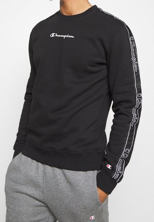 Mann trägt schwarzen Champion-Sweatshirt mit Logo auf Brust und Ärmeln, kombiniert mit grauer Jogginghose mit kleinem Logo am Oberschenkel.