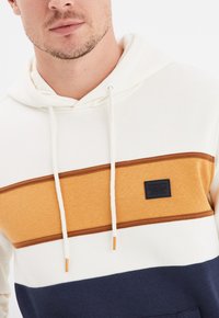 Sudadera con capucha que presenta bloques de color crema, mostaza y marino. Fabricada con un tejido suave, cuenta con un parche de logo en la parte delantera y detalles con cordón.