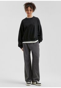 Giovane donna con capelli ricci, che indossa un maglione nero oversize, pantaloni grigi a gamba larga e sneakers bianco e nere, in piedi contro uno sfondo semplice.
