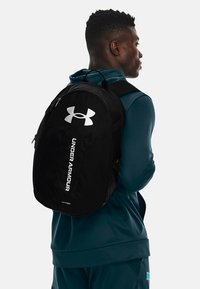 Zaino nero dalla forma arrotondata, con un grande logo bianco Under Armour e testo sul lato, realizzato in tessuto resistente.