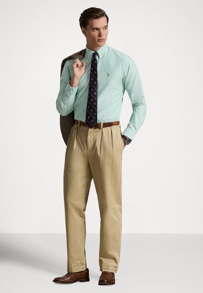 Šviesiai žalia marškinių palaidinė, tamsiai mėlynas kaklaraištis su aukso raštu, smėlio spalvos kelnės, rudi odiniai batai ir pilkas švarkas užmestas per petį.