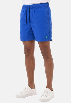 Man wearing bright blue drawstring shorts with a small green logo on the left leg and white lace-up sneakers, standing against a plain background.