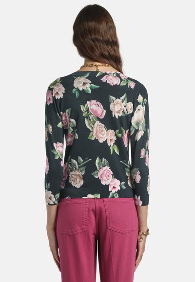 Pull à fleurs vert foncé avec des roses roses et crème. Manches trois-quarts et ourlet côtelé. Associé à un pantalon rose.