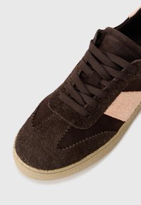 Brune suedé sneakers med en teksturert overdel, rosa aksenter på siden, og beige gummisåle. Har flate lisser og sikksakk-søm.