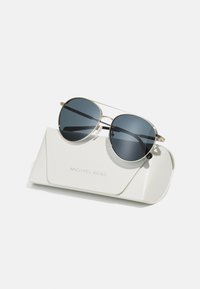 Lunettes de soleil aviateur en métal doré avec des verres foncés reposant sur un étui texturé blanc portant le logo "MICHAEL KORS" en relief.