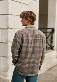 Bruine geruite shirt met blauwe en rode accenten, voorzien van een button-down kraag, lange mouwen en een ontspannen pasvorm, gedragen over blauwe jeans.