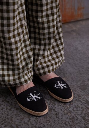 Voeten met zwarte Calvin Klein instapschoenen met touwzolen, gecombineerd met losse groene en witte geruite broeken op een gestructureerde vloer.