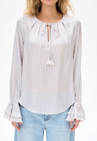 Femme portant un chemisier blanc à manches longues avec des pois bleus, des manches à volants, un col à nouer et un jean bleu clair.