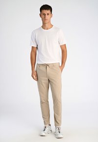 Pantalon en coton beige avec taille élastique, design slim-fit, associé à un t-shirt blanc uni et des baskets blanches avec des lacets colorés.
