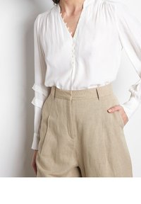 Kvinna iklädd en vit blus med knappar och beigefärgade höga byxor, med ena handen i fickan, mot en enkel vit bakgrund.