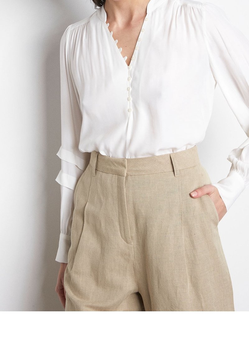 Kvinna iklädd en vit blus med knappar och beigefärgade höga byxor, med ena handen i fickan, mot en enkel vit bakgrund.