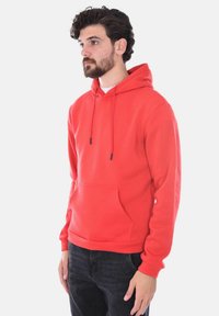 Felpa rossa realizzata in tessuto morbido, con tasca frontale, cappuccio regolabile e polsini a coste. Indossata con jeans scuri.