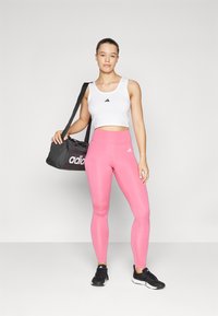 Biely crop top s čiernym logom, ružové legíny, čierne tenisky a čierna športová taška s bielym brandingom. Hladká tkanina, priliehavý strih.