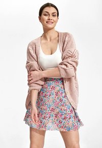 Jasnoróżowy sweter z dzianiny o koronkowym wzorze i podwiniętych r�ękawach, noszony na białym topie na ramiączkach oraz zwiewnej, wielokolorowej spódnicy w kwiaty.