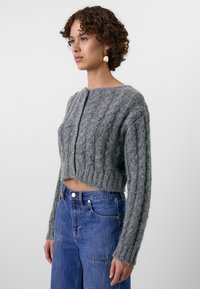 Pull gris, court, en tricot doux et texturé avec un motif en torsades, doté d'un col rond et de boutons dorés sur le devant.