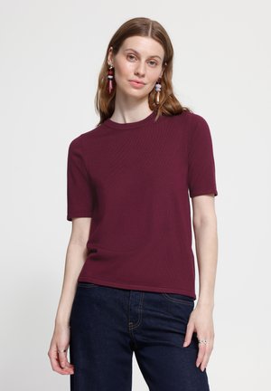 Jeune femme portant un haut bordeaux à manches courtes, un jean foncé et des boucles d'oreilles pendantes colorées, debout devant un fond clair uni.