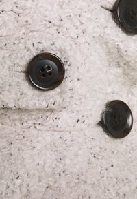 Texturerad ljusrosa tyg med mörka fläckar, som har två stora mörka knappar med fyra hål vardera. Närbild av klädesplaggets material.